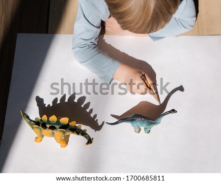 child draws with pencil contrasting shadows from toy dinosaurs. drawing of preschooler, creative ideas for children's creativity. Interesting activities for children during period of self-isolation