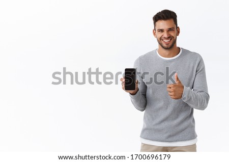 Satisfied handsome bearded male model in grey sweater, show thumb-up and smiling in approval, holding mobile phone, recommend smartphone application, advice download app, white background Royalty-Free Stock Photo #1700696179