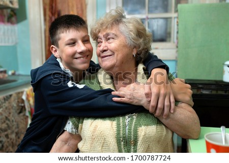 Happy grandmother and grandson hug each other in the kitchen at home. Intergenerational relationships, family values, love, care. Poor, ordinary people. Royalty-Free Stock Photo #1700787724