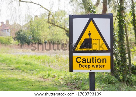 Yellow warning sign with the message of 'Caution, Deep Water' by a village pond.