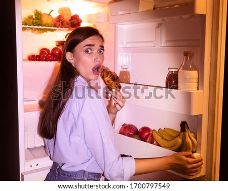Failure of diet. Young woman eating junk croissant near opened fridge late in night, free space Royalty-Free Stock Photo #1700797549