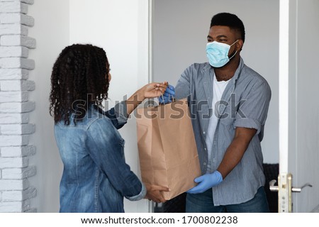 Food delivery during coronavirus. Black courier guy wearing medical mask delivering grocery order to young woman's home