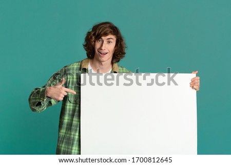 Funky millennial guy pointing at poster with empty space for your ad on turquoise background