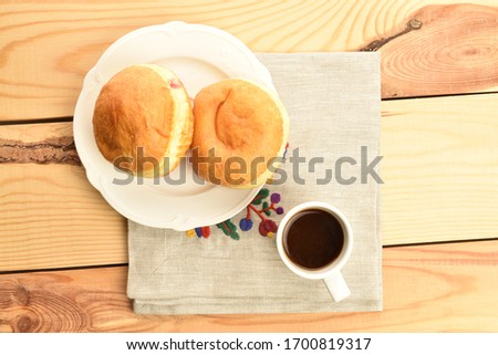 Two freshly baked appetizing, tasty, sweet donuts with cherry filling on a white ceramic saucer with a cup and linen napkin, close-up, on a background of a table made of wood, top view.