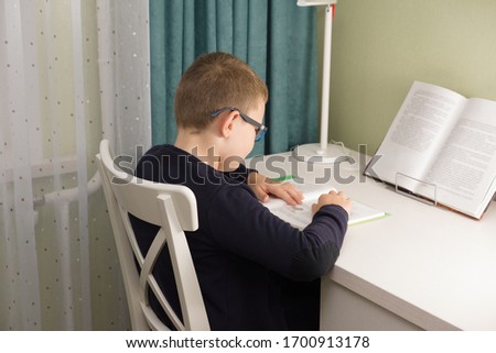 boy in glasses and a jacket sits at a table and does a school task