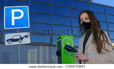 The woman in protective medical mask with the charging cable near the column for charging electic, hybrid cars. Traffic sign - parking and charging electric vehicles. Coronavirus and ecology concept.