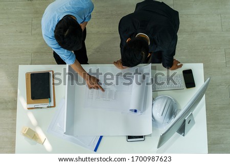 top view photo of asian businessman talked to a young civil engineer and looked at Muck Up  blueprint of building construction plan with  safety hard hat on the table  in the office