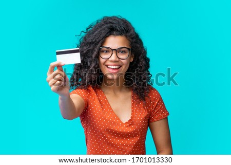 Studio portrait of happy attractive young african american woman in trendy spectacles smiling and holding credit card in hand. Isolated over blue background.