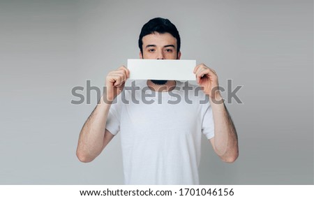 Young man isolated over background. Emotional guy cover face with blank piece of page. Hold it with both hands