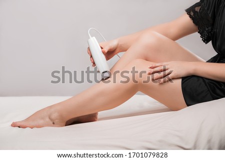 A young woman in a black lace robe does a photo-epilation procedure of her legs on the bed at home, close-up, side view, place for text. Royalty-Free Stock Photo #1701079828