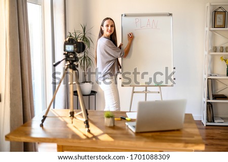 Concept of webinars, online classes, online training. Young woman in casual clothes shows information on a flip chart board and records herself on camera on tripod