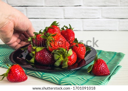 Bright ripe red strawberries in a black plate with green kitchen towel close up on a white brick background with copy space and a human hand