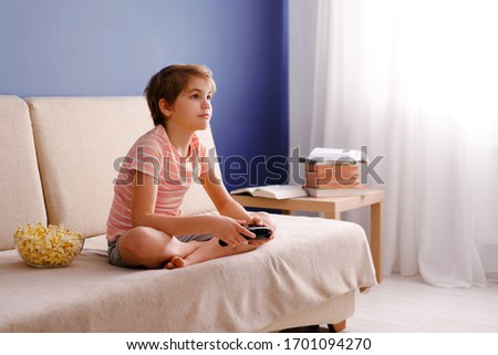 Happy smiling school boy in pink t-shirt sitting on light couch, playing video games with a gamepad instead of doing his homework at home and eating pop corn. Blue background and free space for text.