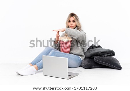 Teenager blonde girl eating popcorn while watching a movie on the laptop making time out gesture