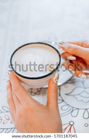 in the foreground, the girl's hands are holding a Cup of coffee cappuccino in the background, a table and a plate, the background is blurred