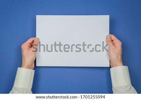 Mans hand hold a sheet of white paper on the royal blue background. Place for a text.