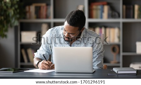 Concentrated African American male worker sit at desk handwrite watching webinar or training on laptop, focused biracial man make notes busy studying working on computer at workplace in office Royalty-Free Stock Photo #1701296191