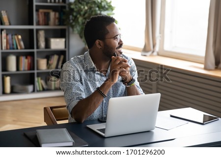 Smiling African American man in glasses sit at desk distracted from computer work look in distance visualizing, happy biracial male lost in thoughts dreaming or thinking, business vision concept Royalty-Free Stock Photo #1701296200