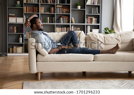 Smiling young African American man relax on comfortable couch in living room using modern laptop, happy millennial biracial male rest on cozy sofa at home working or studying on computer gadget Royalty-Free Stock Photo #1701296233