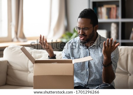 Unhappy African American man client disappointed with product quality shopping online, mad biracial male open cardboard package frustrated with wrong Internet order, bad delivery service concept Royalty-Free Stock Photo #1701296266