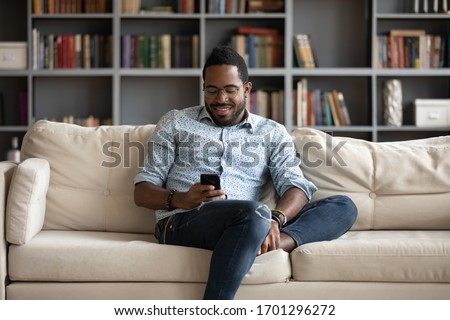 Smiling african American man sit relax on comfortable couch at home using modern cellphone, happy biracial millennial male rest on sofa in living room texting messaging on smartphone gadget Royalty-Free Stock Photo #1701296272