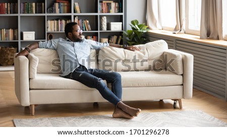 Calm African American young man sit relax on comfortable couch in living room look in distance thinking, relaxed biracial millennial male rest on sofa at home, breathe fresh air, stress free concept Royalty-Free Stock Photo #1701296278
