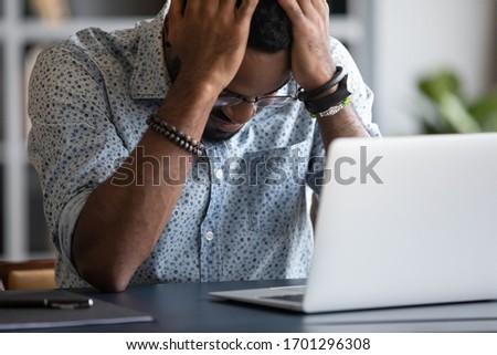 Stressed african American male employee sit at desk feel distressed with bad negative news online, frustrated biracial man student work on laptop having problems, miss deadline or appointment Royalty-Free Stock Photo #1701296308