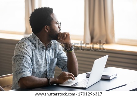 Thoughtful african American male employee in glasses sit at desk distracted from computer work pondering, pensive biracial man look in distance thinking or planning, business vision concept Royalty-Free Stock Photo #1701296317