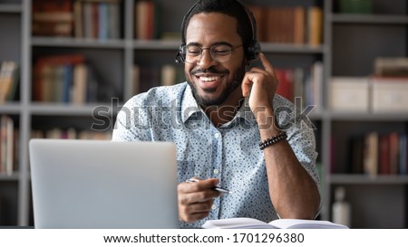 Smiling African American man in glasses and headset watch webinar on laptop making notes, happy biracial male student worker in headphones handwriting studying or working using computer Royalty-Free Stock Photo #1701296380