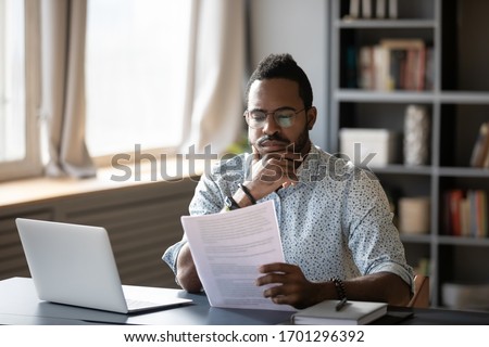 Pensive African American man sit at desk working on laptop reading paper document thinking or analyzing, thoughtful biracial male worker consider paperwork agreement at office or home workplace Royalty-Free Stock Photo #1701296392