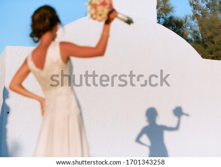 A dark-haired bride with a bouquet in her hand stands in front of a white wall with her shadow.