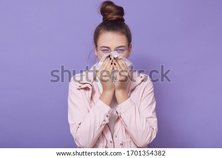 Indoor picture of sick upset cute young lady holding handkerchief in hands, blowing her nose, having caught cold, feeling unwell, having bun, wearing trendy eyeglasses. Health problems concept.