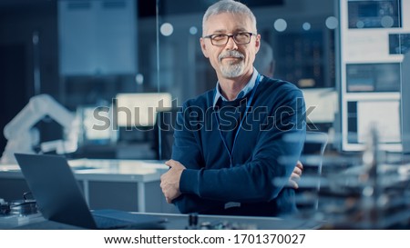 Professional Electronics Design Engineer Wearing Glasses Works on Laptop Computer in Research Laboratory. In the Background Motherboards, Circuit Board, Heavy Industry Robotic Components Royalty-Free Stock Photo #1701370027