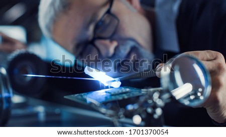 Close-up Portrait of Focused Middle Aged Engineer in Glasses Working with High Precision Laser Equipment, Using Lenses and Optics for Accuracy Electronics. Testing Superconductor Material Royalty-Free Stock Photo #1701370054