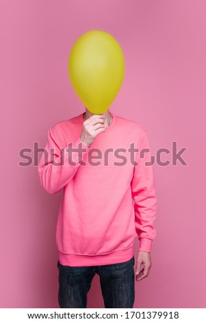 Vertical picture of tall man in pink sweater hide face behind green balloon. Stand alone and pose in studio. Isolated over pink background