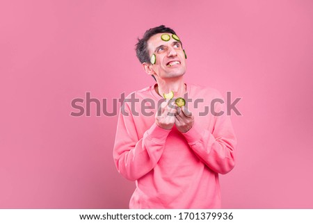 Picture of young man with evil emotions on his face look up. Hold cucumber pieces in hands and on face. Isolated over pink background