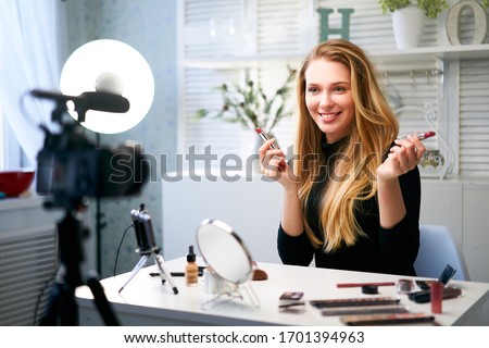 Beauty blogger woman filming daily make-up routine tutorial near camera on tripod. Influencer blonde girl live streaming cosmetics product review in home studio. Vlogger female comparing lipsticks. Royalty-Free Stock Photo #1701394963