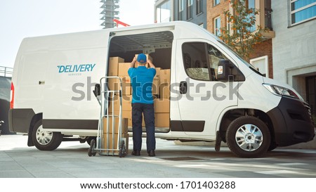 Delivery Man Uses Hand Truck Trolley Full of Cardboard Boxes and Packages, Loads Parcels into Truck / Van. Professional Courier / Loader helping you Move, Delivering Your Purchased Items Efficiently Royalty-Free Stock Photo #1701403288