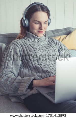 Young freckled woman is siting on sofa in a modern scandinavian apartment. She fulfilling her free time at home with digital entertainment while having a video call with friends. Stay sociable. 