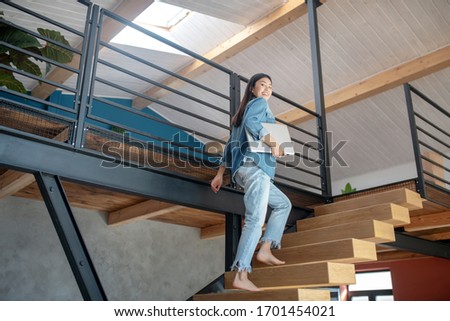 At home. Young woman going upstairs, holding laptop under arm, smiling Royalty-Free Stock Photo #1701454021