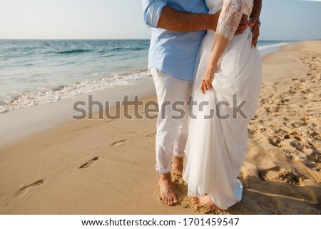Newlyweds honeymoon. Barefoot bride in white dress and groom in white pants walking on romantic ocean beach. Young couple foots on golden sand.