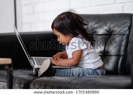 Side view of talented little mixed race girl pushing button on laptop while looking at cartoons while lying on an armchair. Concept of independent children and gadgets. Children and internet addiction