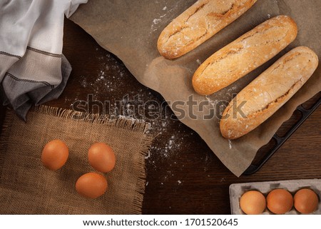 Fresh baguettes from the oven on a baking sheet on a brown wooden kitchen table on which flour is scattered, as well as a tablecloth on the left and eggs in a cardboard box and on a burlap