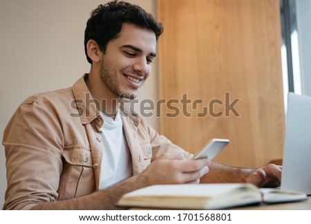 Indian freelancer holding smartphone, communication, working from home. Handsome asian man using laptop computer, internet, planning project, sitting in modern office 