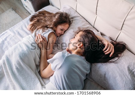 young mother and daughter woke up in the morning in a bright room, they lie in bed looking at each other, mother stroking her daughter