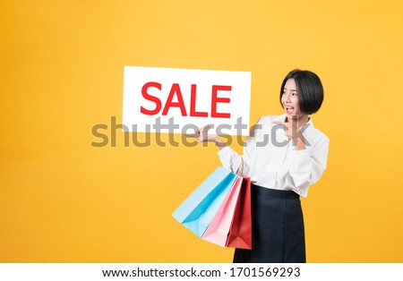 Young smiling asian woman holding multi coloured shopping bags and hands showing sale sign on light orange background.