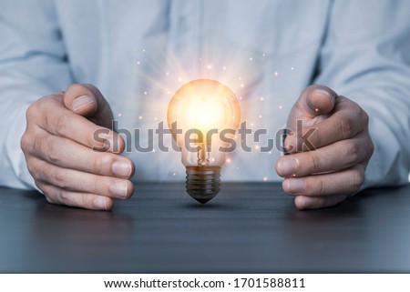 Two hands protecting the light bulb that is illuminating on the table. Creative protecting patents and ideas concept. Royalty-Free Stock Photo #1701588811