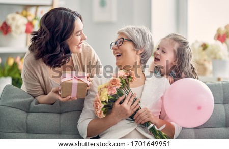 Happy mother's day! Child daughter is congratulating mom and granny giving them flowers and gift. Grandma, mum and girl smiling and hugging. Family holiday and togetherness.  