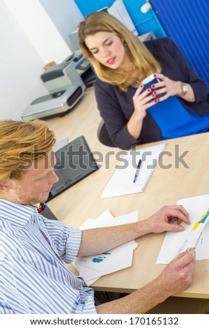 Young designer showing his work to a female coworker, sitting behind a large desk in a design studio, discussing product ideas and conceptual sketches. Focus on the man in front