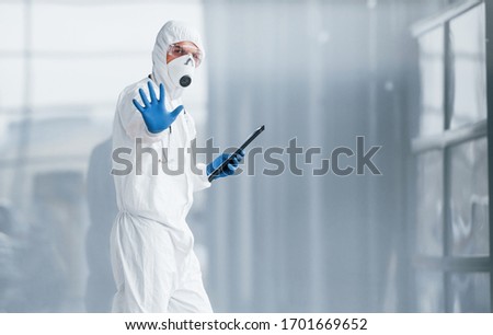 Shows stop gesture by hand. Male doctor scientist in lab coat, defensive eyewear and mask holding notepad in hands.